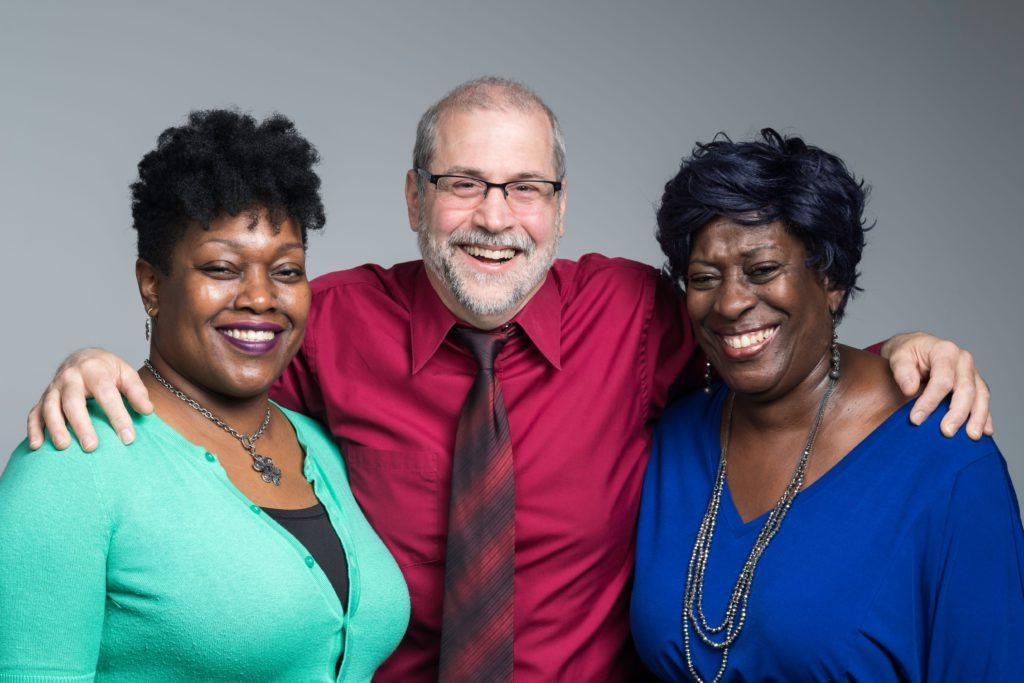 Three individuals dressed in business attire, smiling with their arms wrapped around each other's shoulders