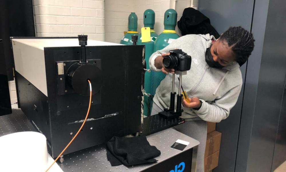 High school student in a mask focuses a camera near a spectrometer.