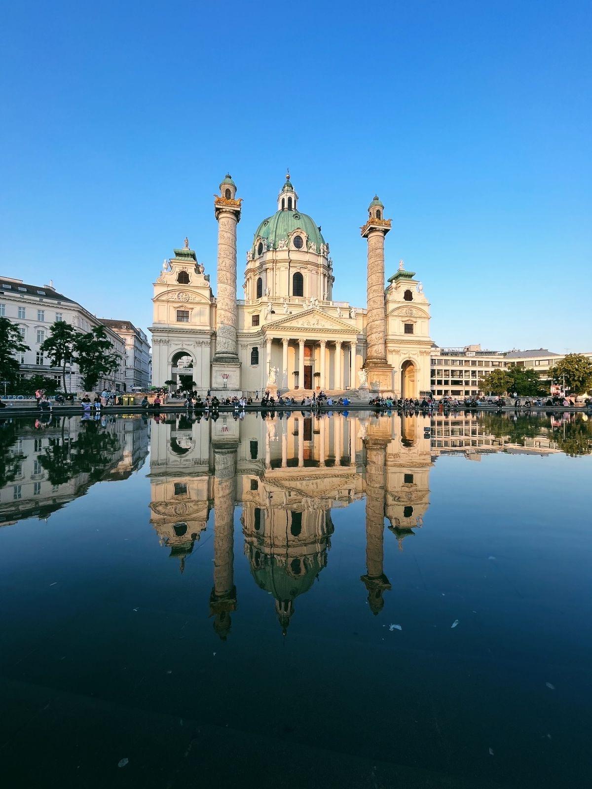 A grand baroque-style white cathedral with a large green dome and twin columns stands against a clear blue sky. 这座建筑在宁静中映出美丽的倒影, glassy pool of water in the foreground, with people gathered around its base.