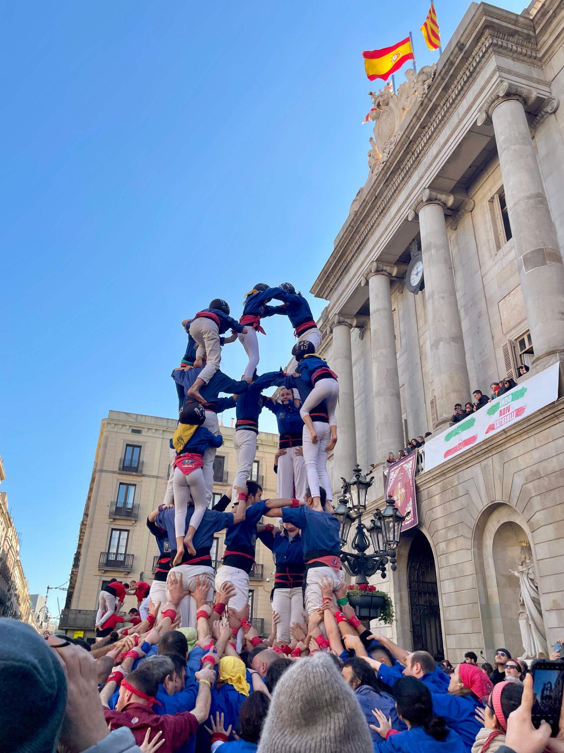 A human tower is formed by a group of people, standing in front of a historic building with columns. Flags are seen atop the building. The base of the tower is wide, gradually narrowing as it reaches the top, with several tiers of participants. Onlookers are gathered around.