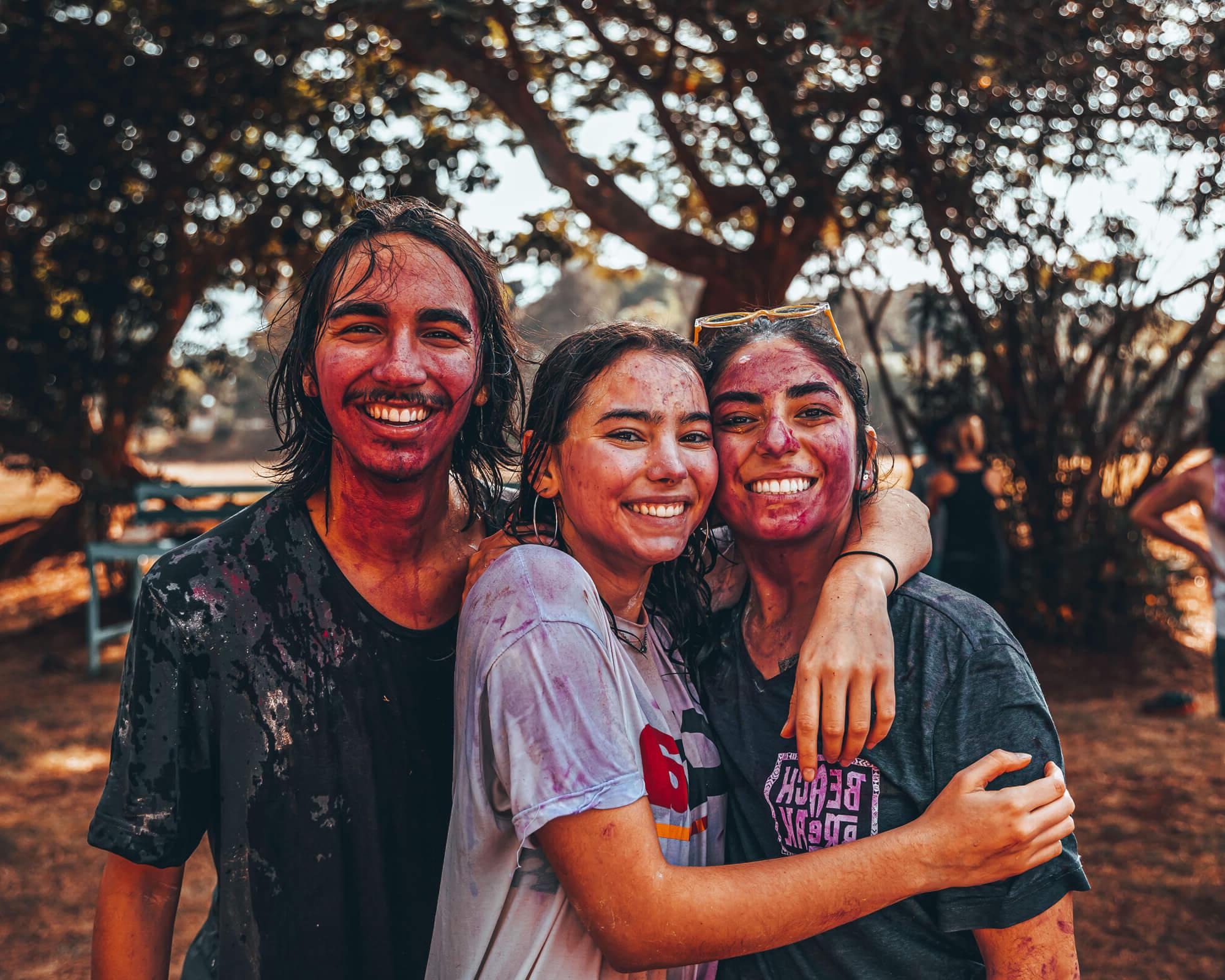 三个年轻人站在外面，身上涂满了五颜六色的粉末, smiling and hugging, with trees in the background.