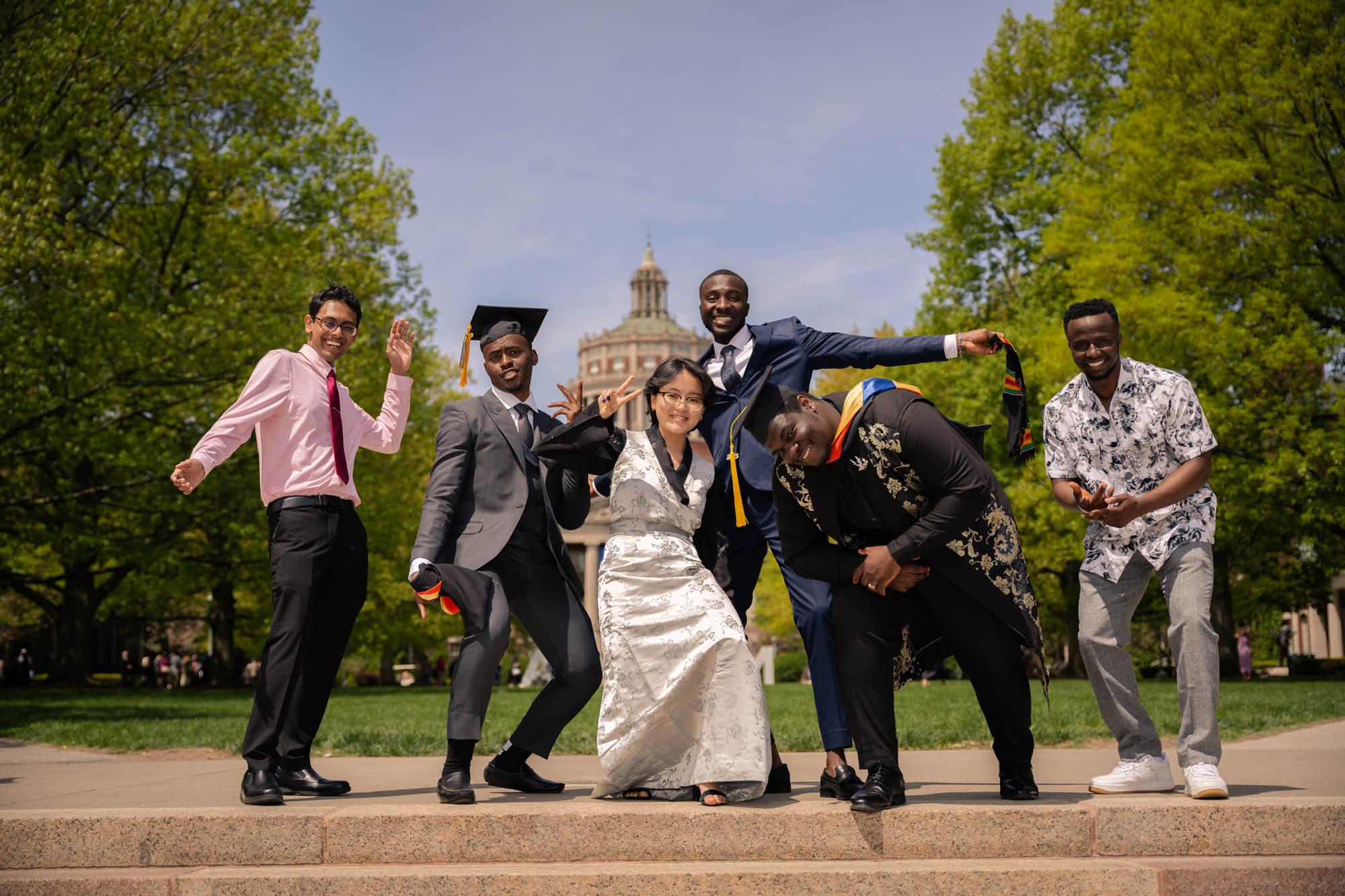 A group of six diverse graduates, dressed in caps, gowns, and formal wear, joyfully pose for a celebratory photo on a sunny day in front of a historic building surrounded by lush green trees. 他们微笑着，摆着好玩的姿势，流露出兴奋和自豪.